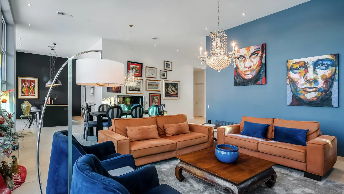 Bright living room with orange sofas, blue chairs, and face art on a blue wall. A chandelier hangs above.