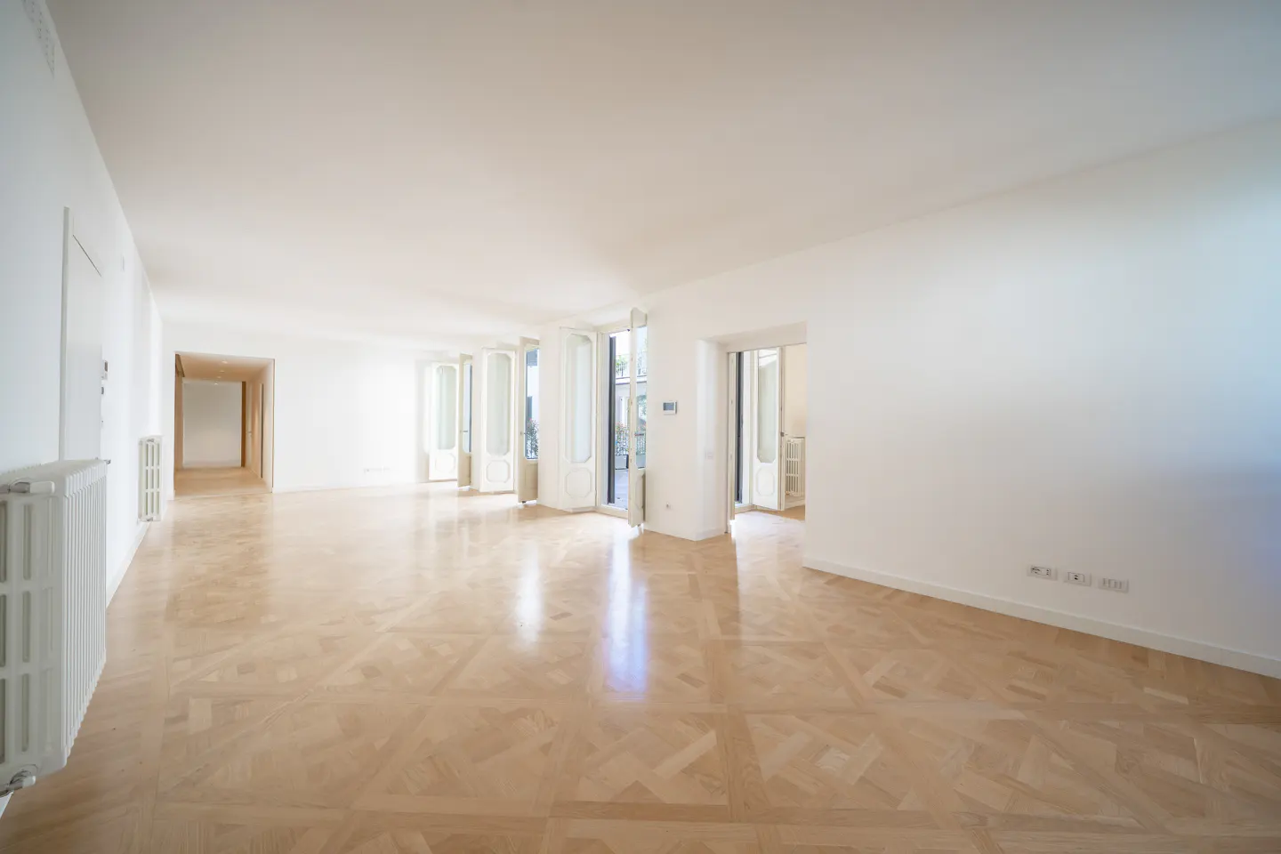 Bright, empty room with white walls, light wood floors, and open white French doors leading to a balcony.