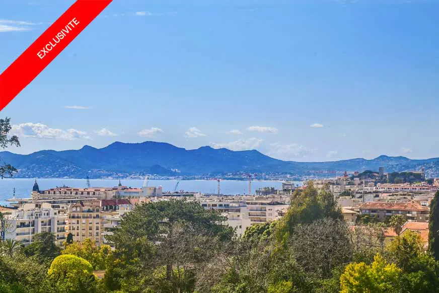 Cityscape view with buildings, trees, and mountains in the background under a blue sky. A red banner with "EXCLUSIVITE" is in the upper left corner.