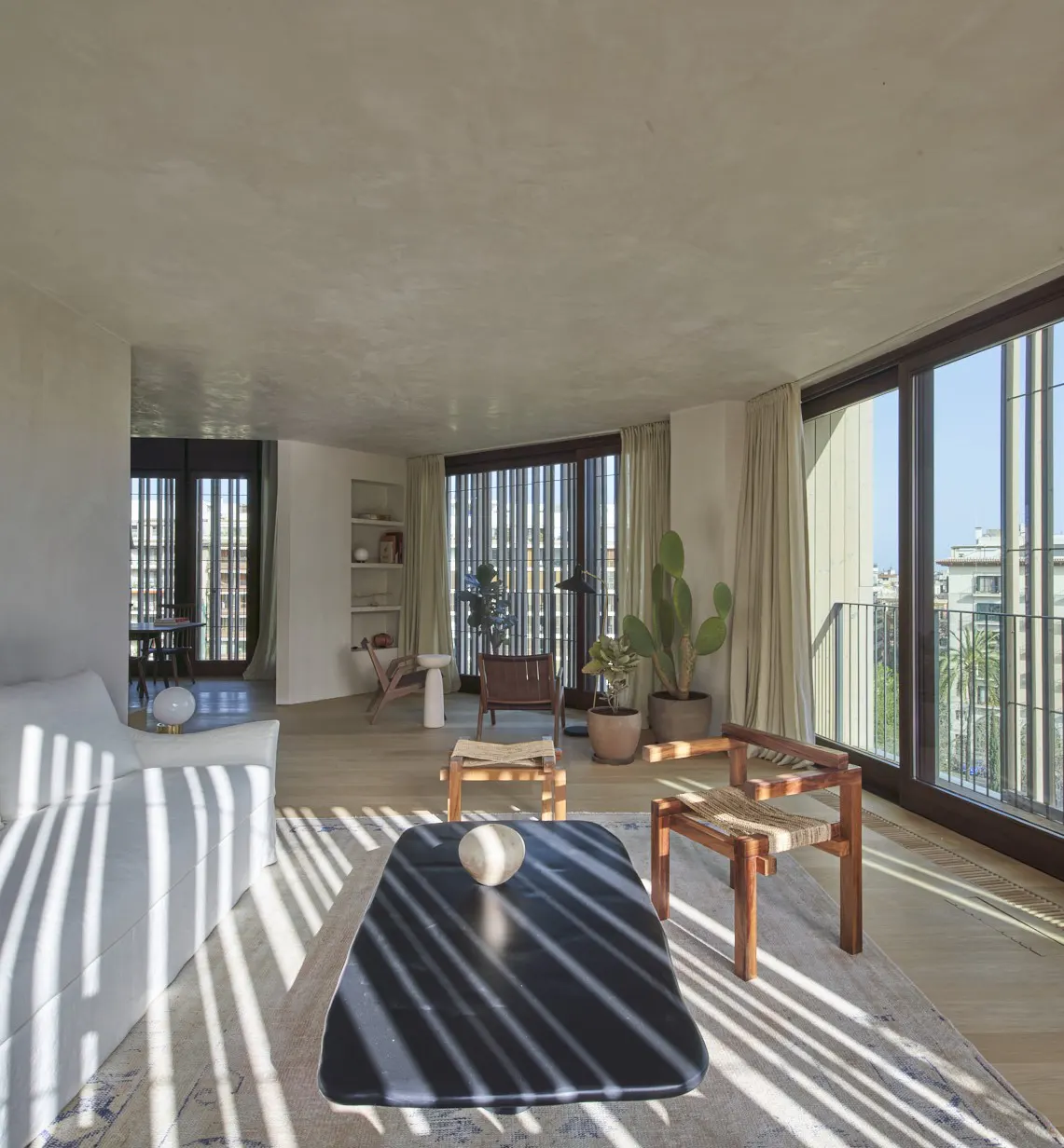 Bright, modern living room with a white sofa, dark coffee table, and wooden chairs. Sunlight streams through the window blinds.