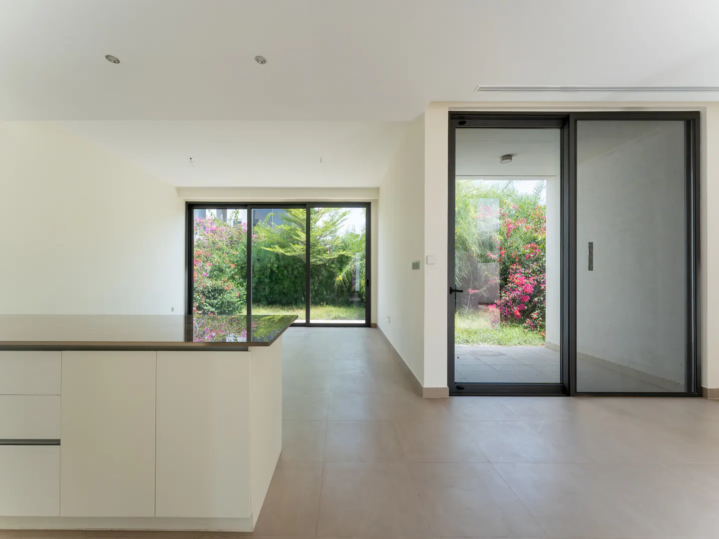 Bright, modern interior with white walls, tile floors, and a kitchen island. Large windows and sliding glass doors offer views of lush greenery.