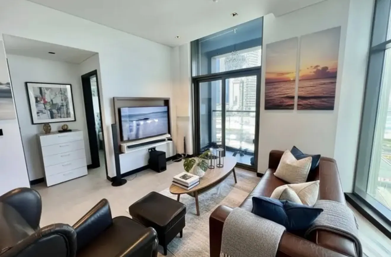 Bright living room with brown leather sofa, black chair, and TV. Large windows offer city views. Artwork and decor add a modern touch.