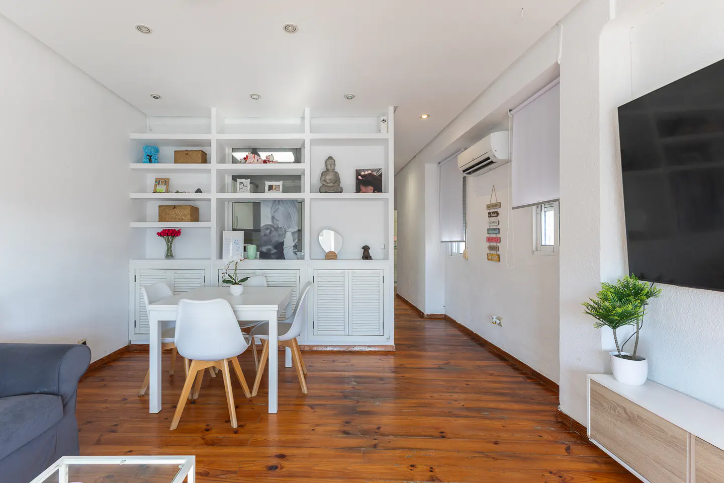 Bright living room with white walls, wood floors, and a white dining set. A large white bookshelf displays decorative items. A TV is mounted on the wall.