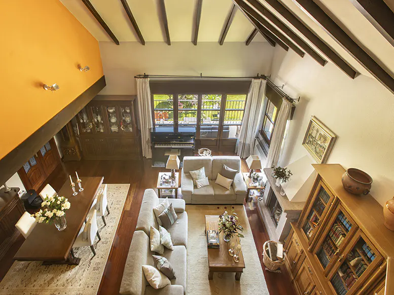 A high-angle view of a living room with wood floors, sofas, a dining table, and a piano. The walls are orange and white, and the ceiling has exposed beams.