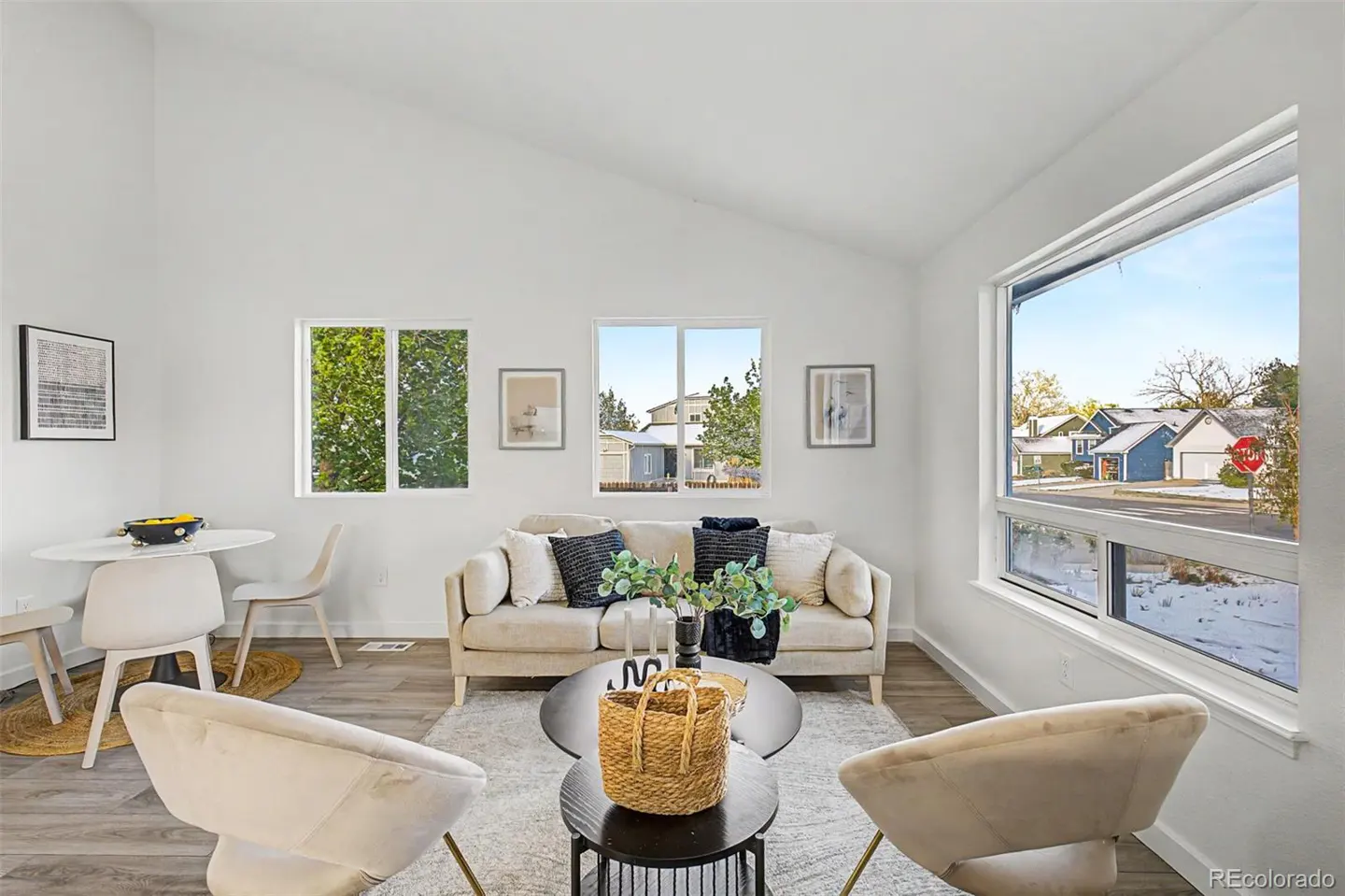 Bright living room with white walls, wood floors, and vaulted ceiling. Beige sofa with pillows, round tables, and two chairs face the camera.