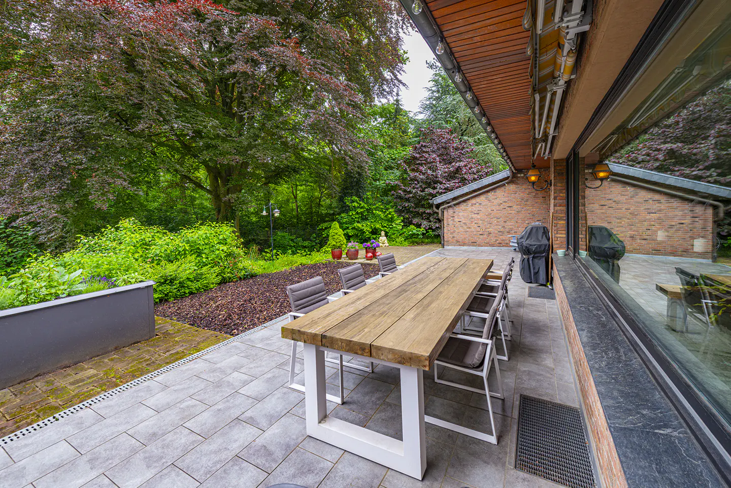 Outdoor patio with a long wooden table and chairs, a grill, and lush green trees in the background.