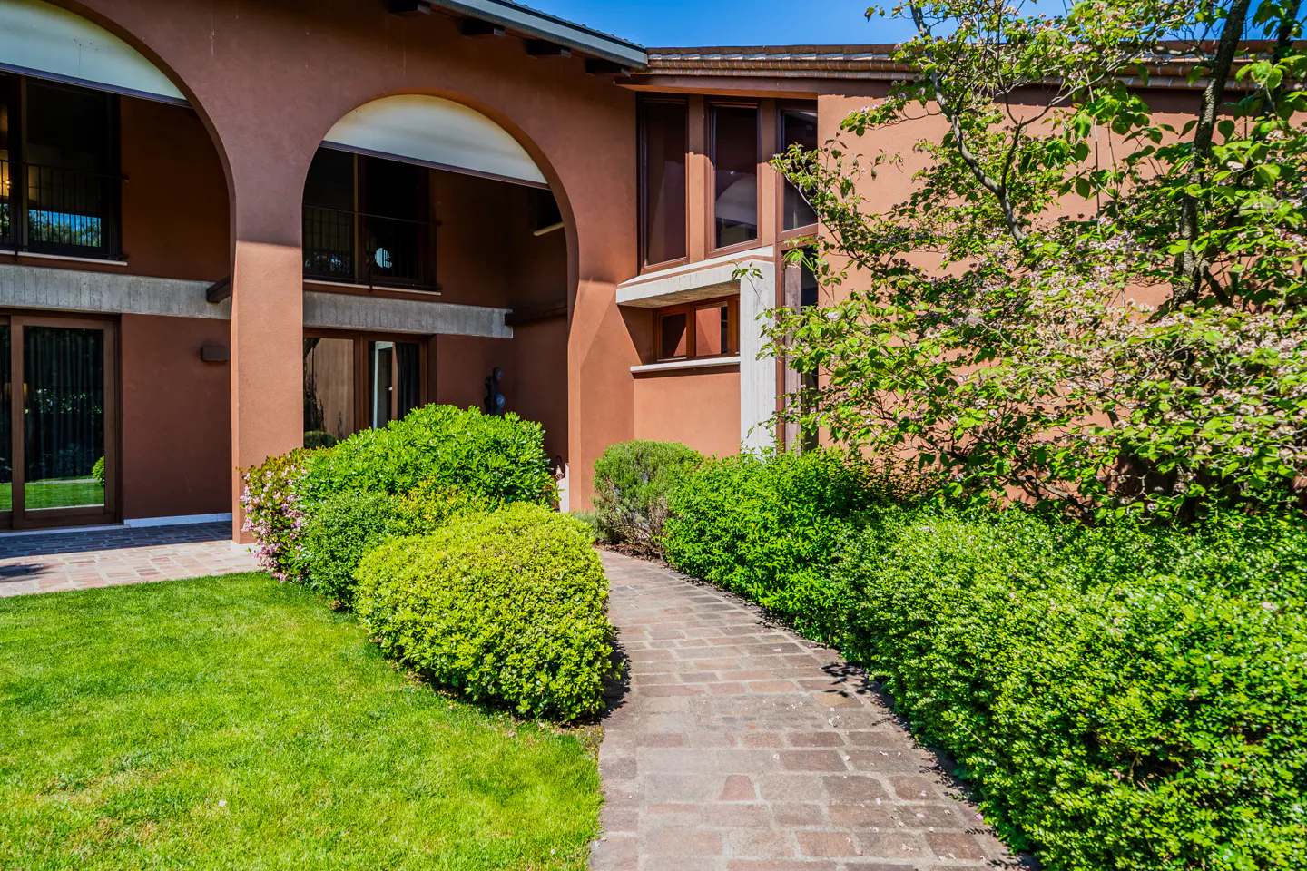 Exterior of a terracotta-colored house with arched entryways, a brick path, green bushes, and a lawn.