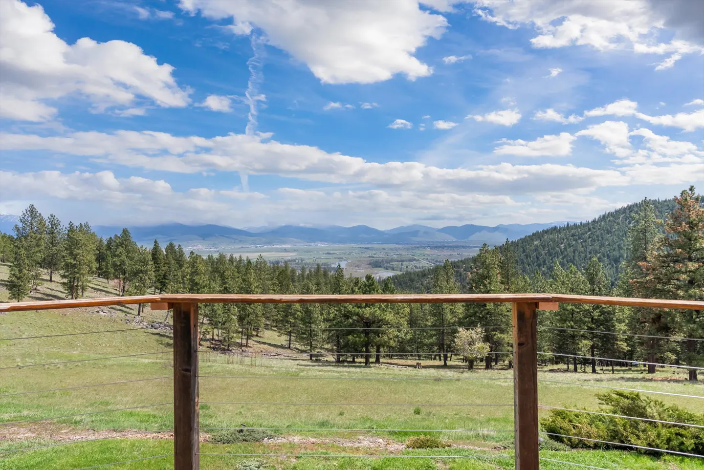 Scenic view from a wooden deck overlooking green hills, pine trees, and distant mountains under a blue sky with white clouds.