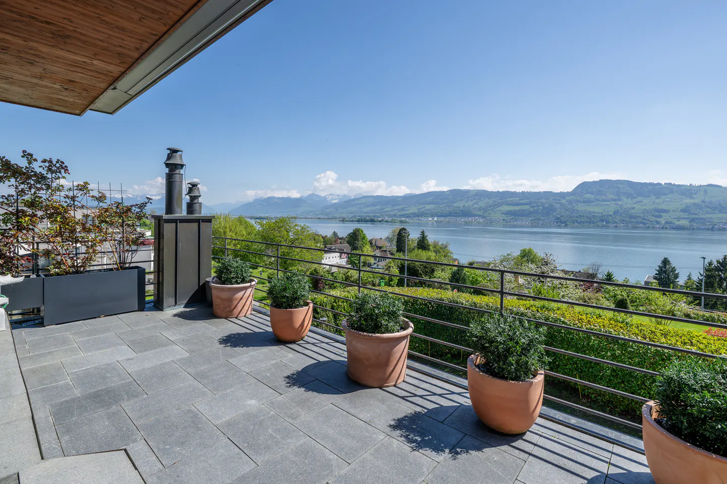 Balcony view with gray stone tiles, potted plants, and a metal railing overlooking a lake and mountains under a blue sky.