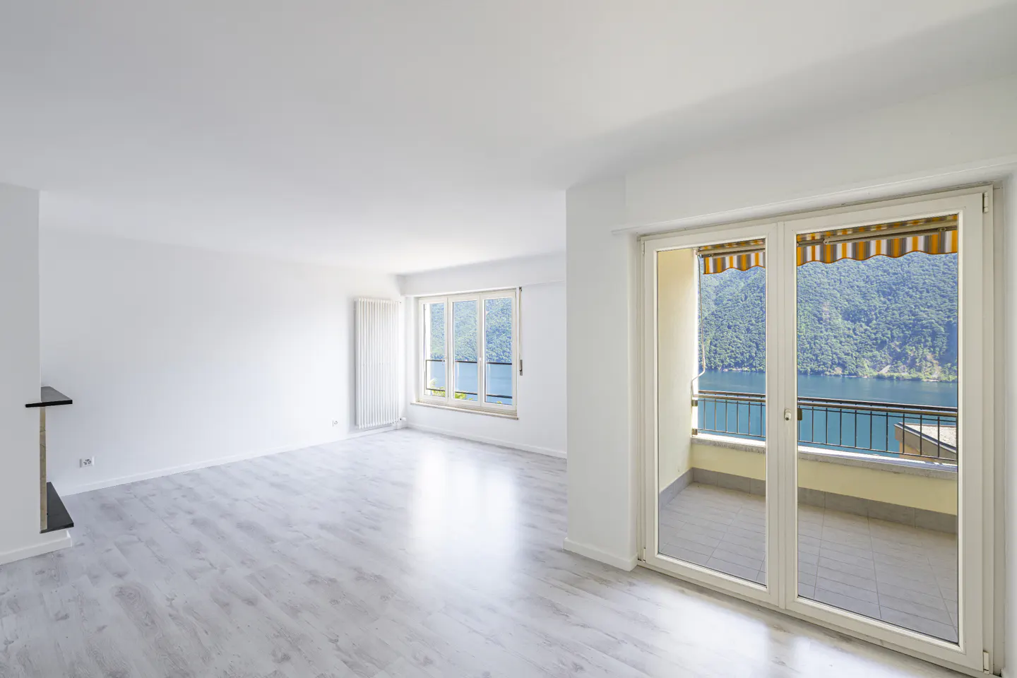 Bright, empty room with light wood floors, white walls, and glass doors opening to a balcony overlooking a lake and hills.