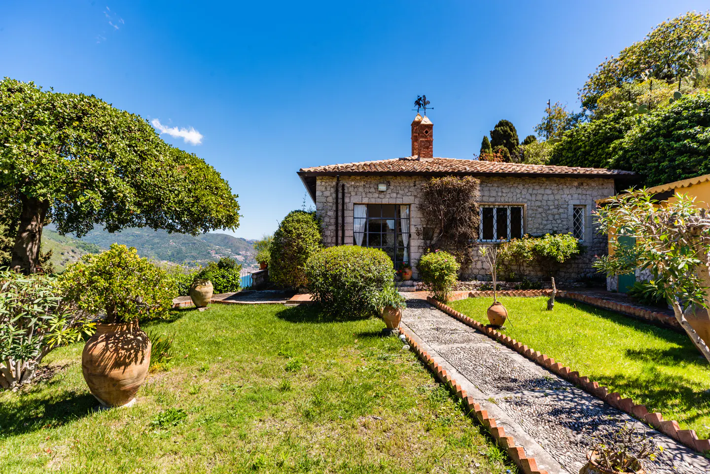 Stone house with a red tile roof, chimney, and weather vane, surrounded by green lawn, trees, and a stone path on a sunny day.