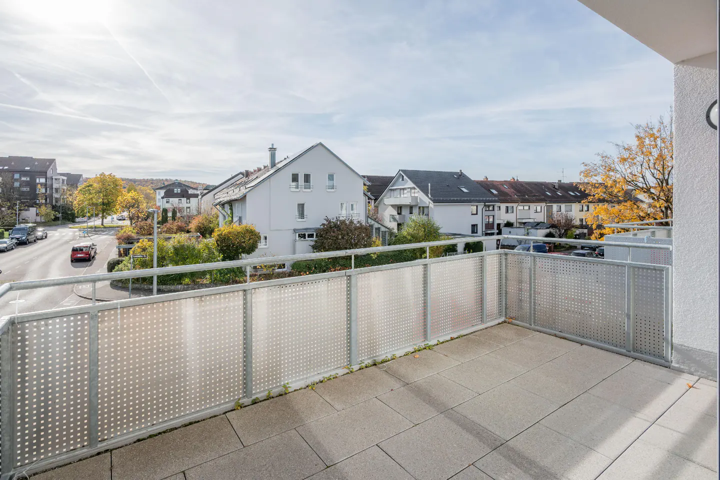 View from a balcony with a gray tiled floor and metal railing, overlooking a street with houses and trees.