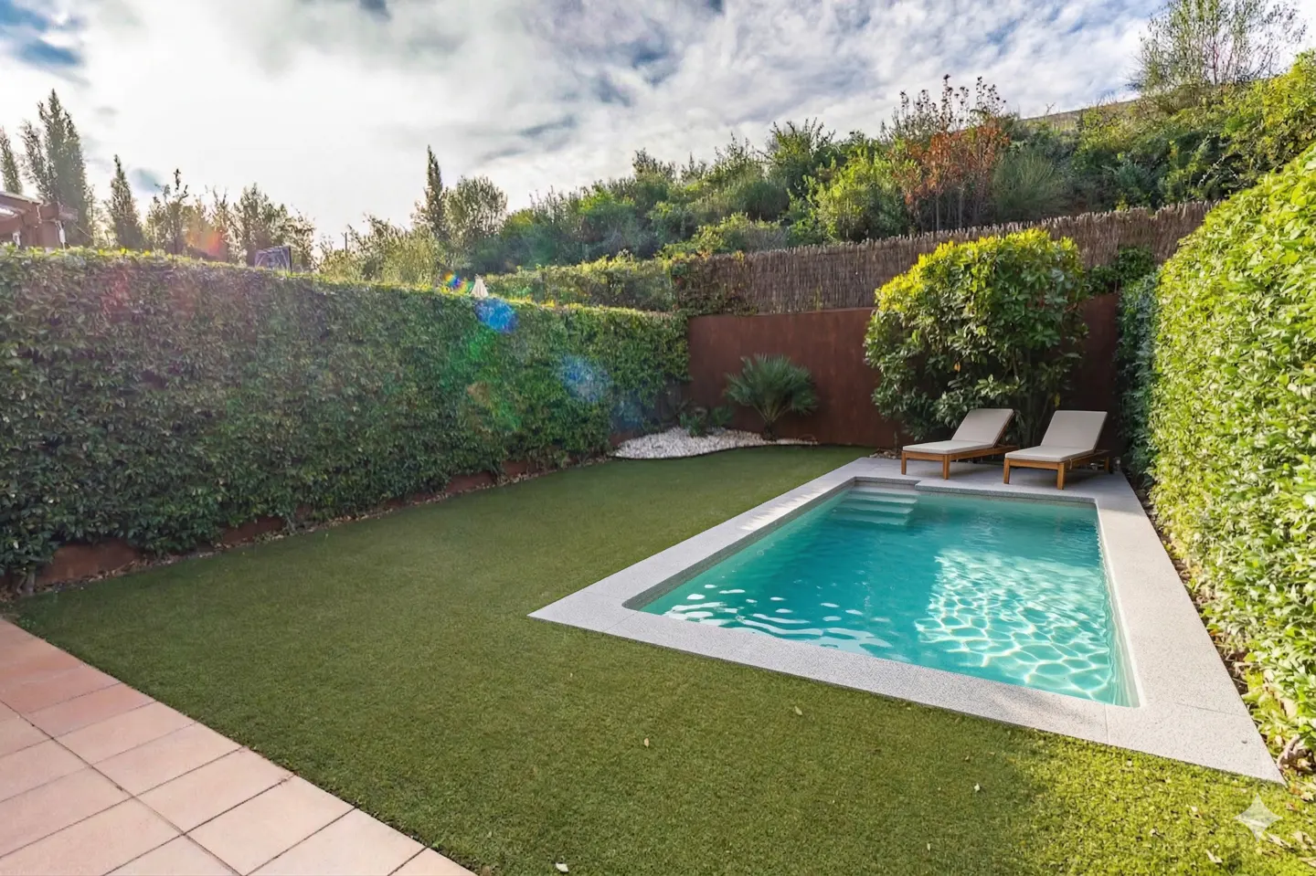Backyard with a rectangular pool, two lounge chairs, green grass, and tall green hedges.
