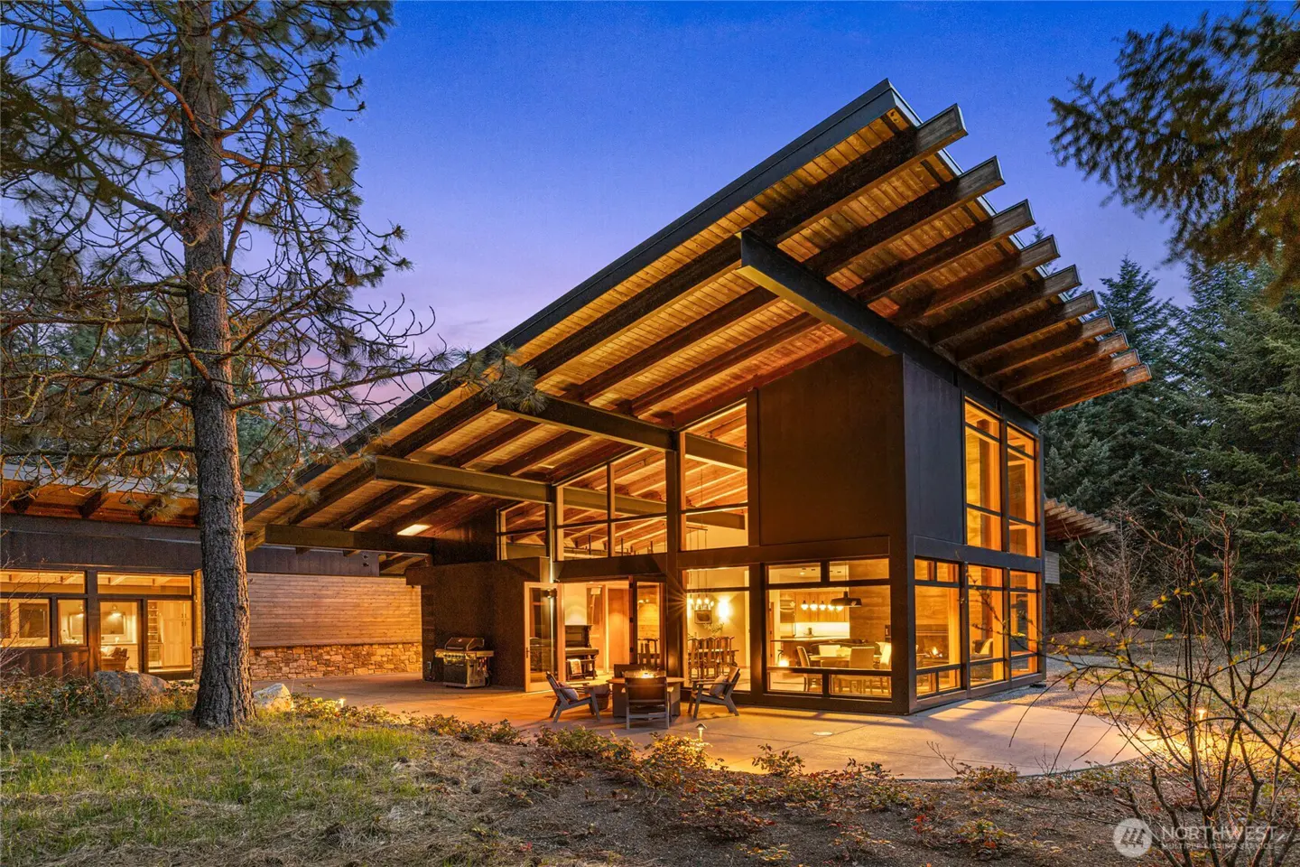 Exterior view of a modern home with large windows, a wooden roof, and a patio with a fire pit at dusk.
