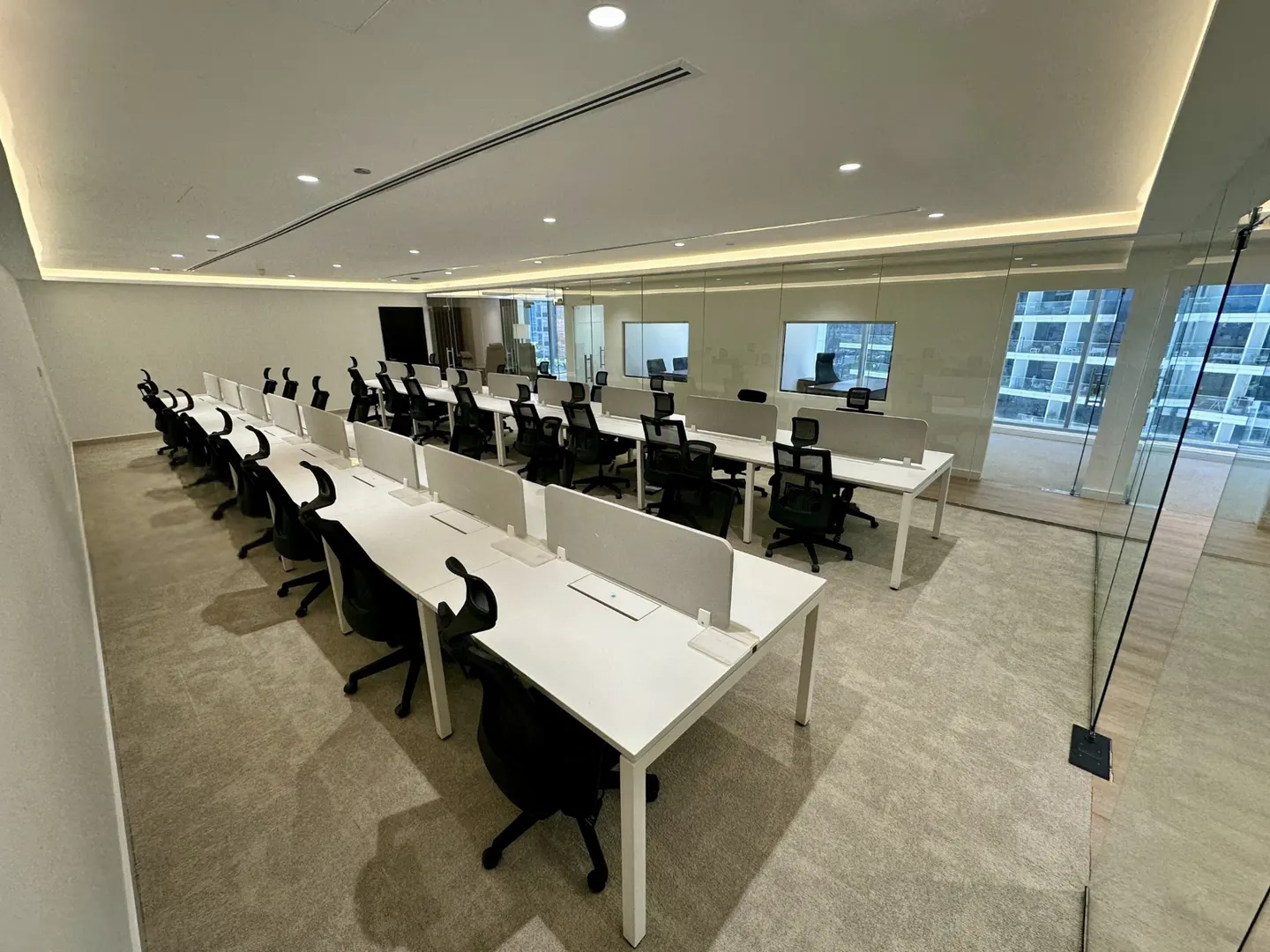 Bright office space with rows of white desks and black chairs. Glass walls offer city views.
