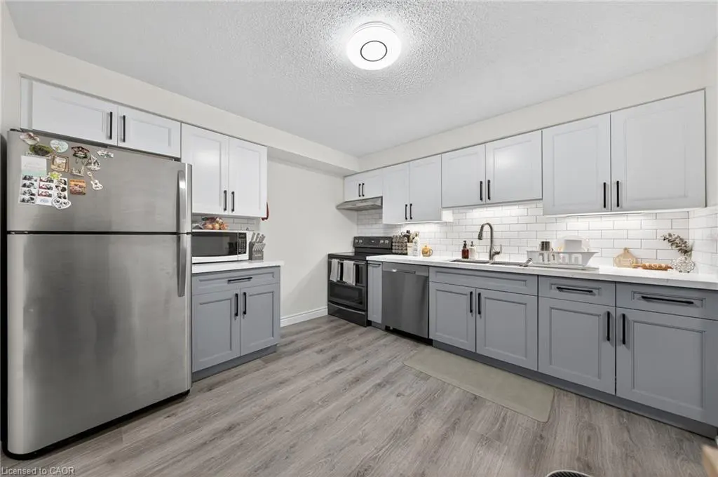 A bright kitchen with gray wood floors, gray lower cabinets, and white upper cabinets. A stainless steel refrigerator is on the left.