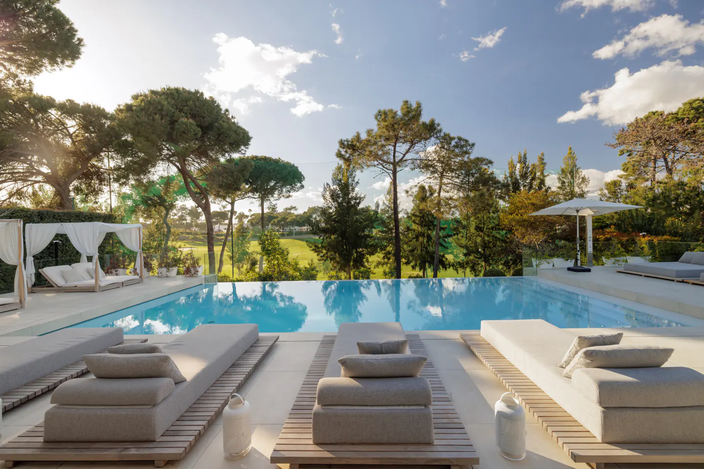 Luxury pool with lounge chairs overlooking a golf course. Blue water reflects the sky. Trees and white cabanas surround the pool.