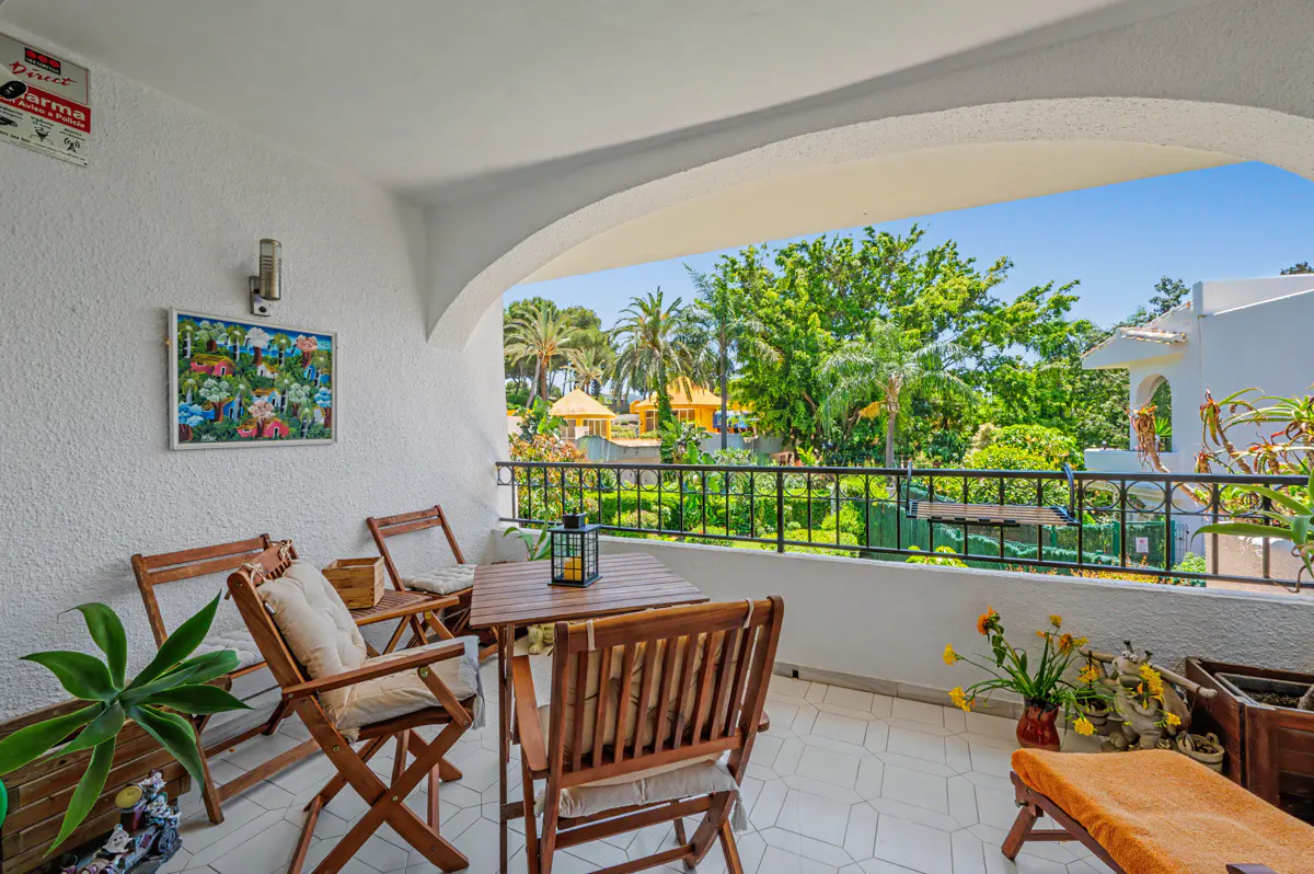 A sunny balcony with a table, chairs, and plants overlooks a lush green garden with palm trees.