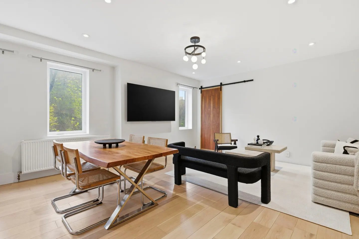 Bright, modern living space with wood floors, a dining table, black bench, and a sliding barn door. A large TV hangs on the white wall.