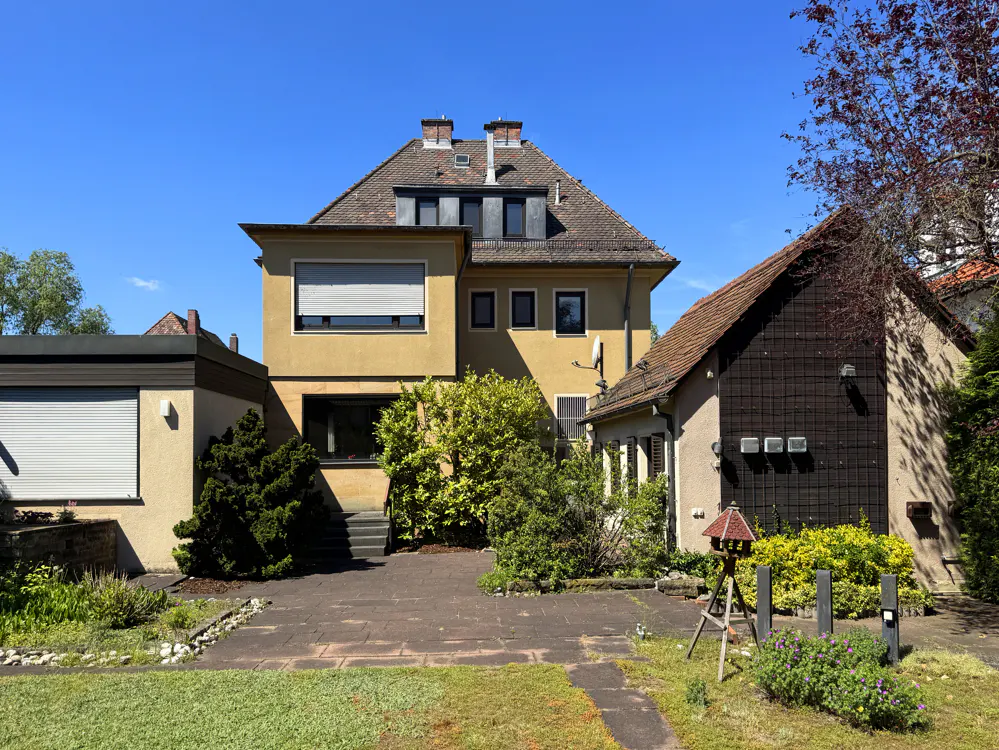 Exterior view of a two-story yellow house with a brown roof, surrounded by green trees and a stone patio.