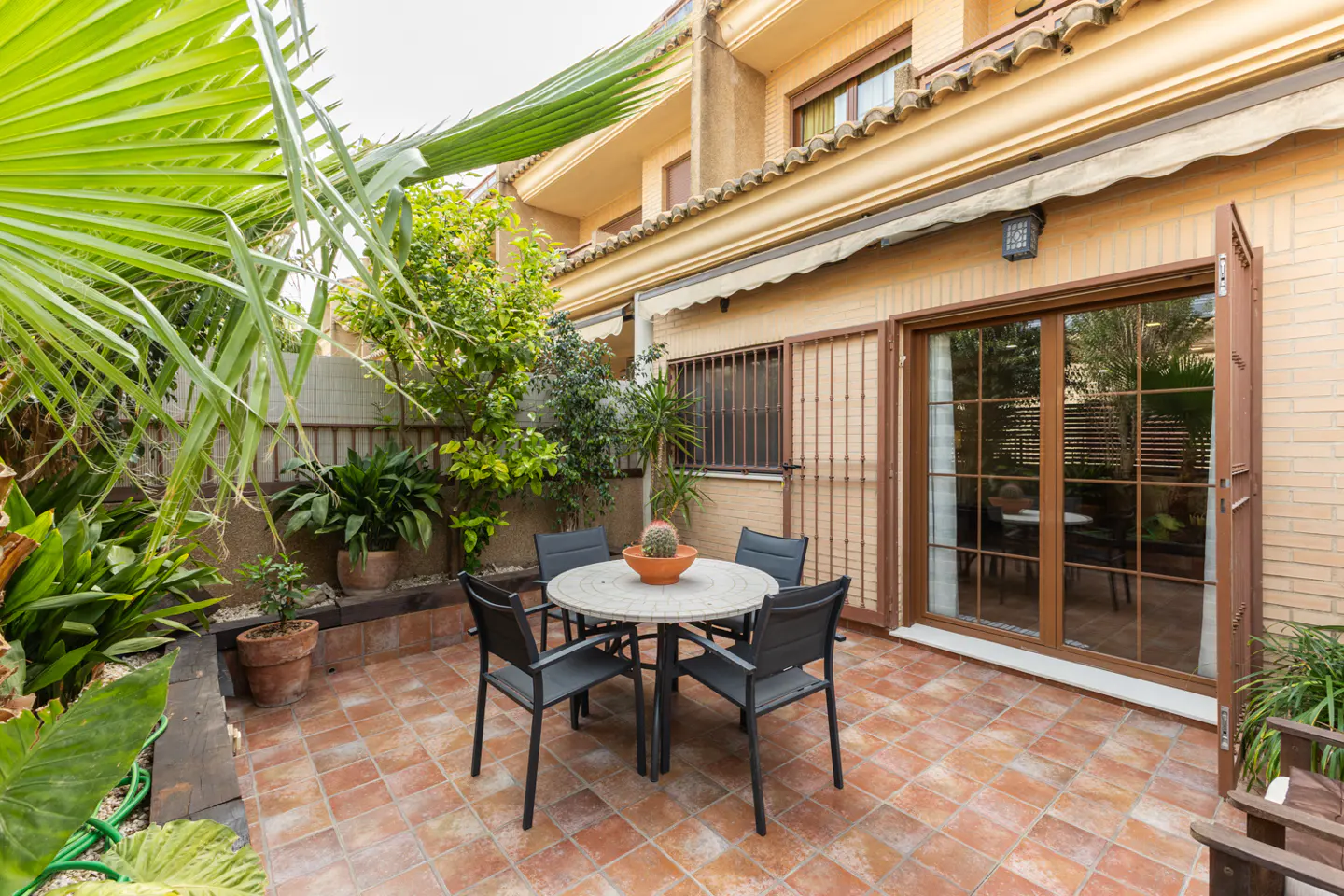Outdoor patio with a round table, four chairs, and lush green plants. Sliding glass doors lead into a tan brick building.