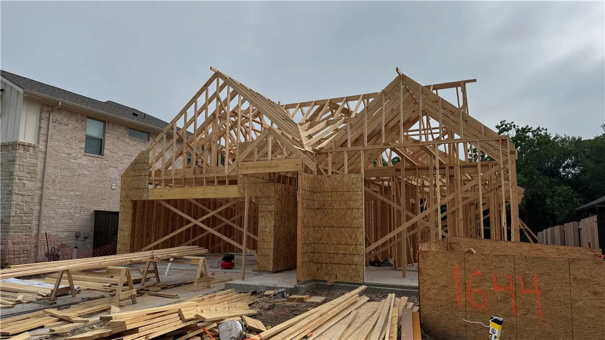 New home construction shows a wood frame and plywood walls. Lumber is stacked on the ground. A brick house is in the background.