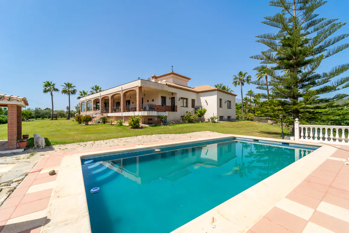 A tan villa with a red tile roof overlooks a turquoise pool on a sunny day. Palm trees and a green lawn surround the property.