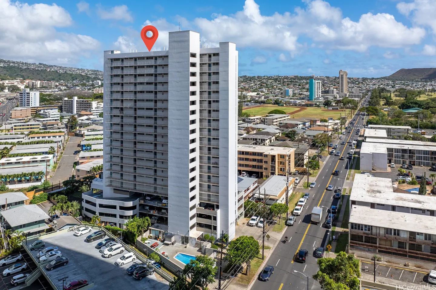 Aerial view of a tall, white apartment building with a red location pin on top, near a busy street and parking lot.