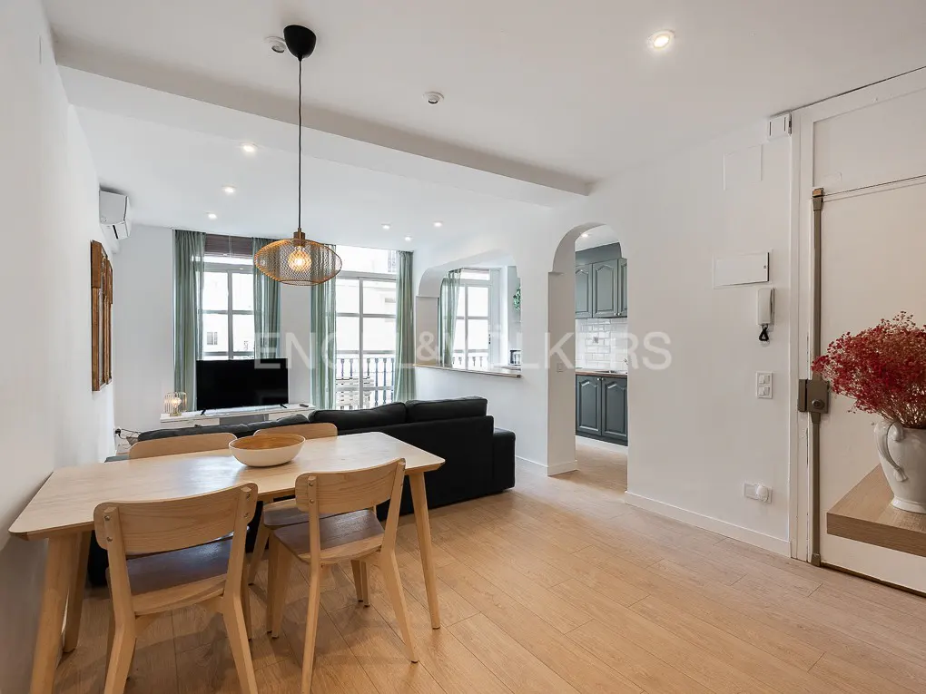 Bright apartment interior with wood floors, a dining table with chairs, a black sofa, and a view into the kitchen.