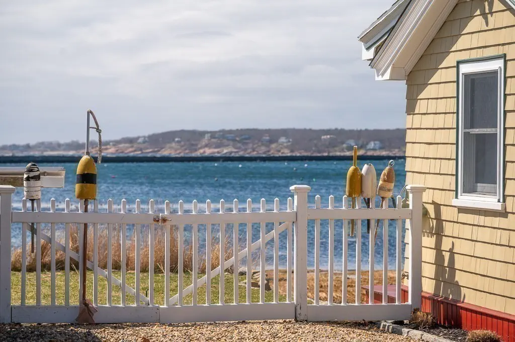 Beach house with a white picket fence adorned with colorful buoys, overlooking the ocean. The house is yellow with white trim.