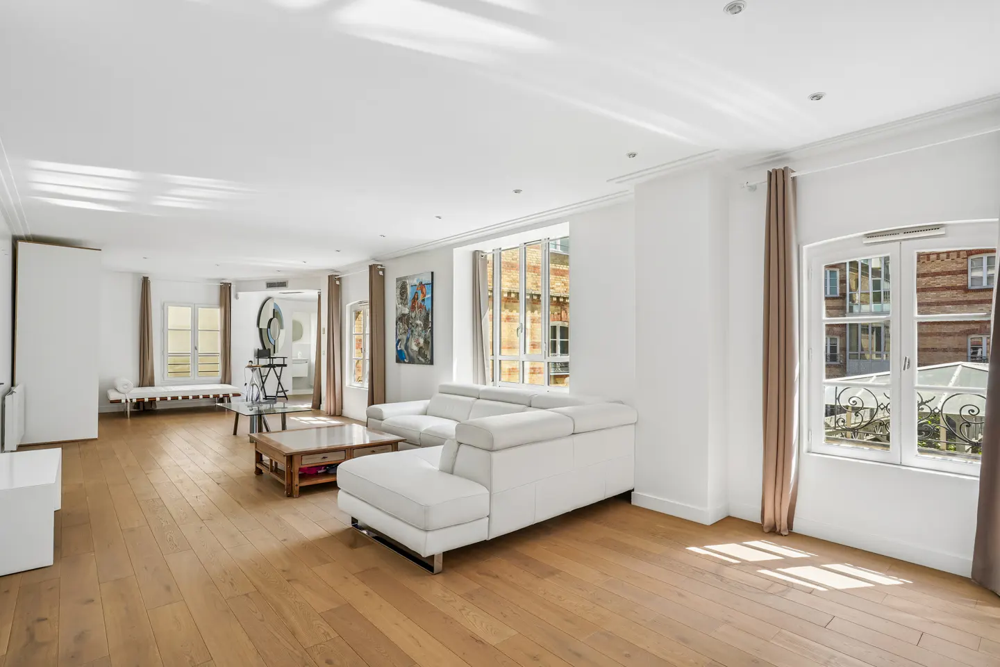 Bright, airy living room with hardwood floors, white walls, and a white sectional sofa. Windows offer city views.