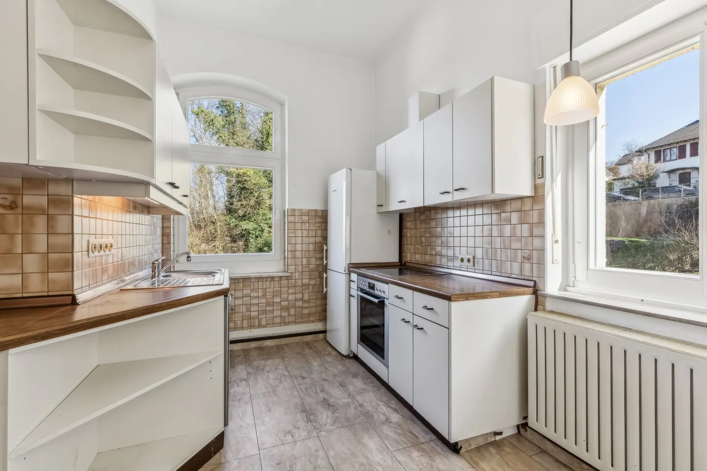 Bright kitchen with white cabinets, brown countertops, and tiled backsplash. A window overlooks trees and a house. A white refrigerator stands next to the oven.