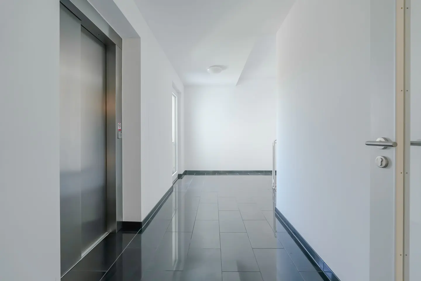 Hallway with black tile floor, white walls, and a stainless steel elevator on the left. A door is visible on the right.