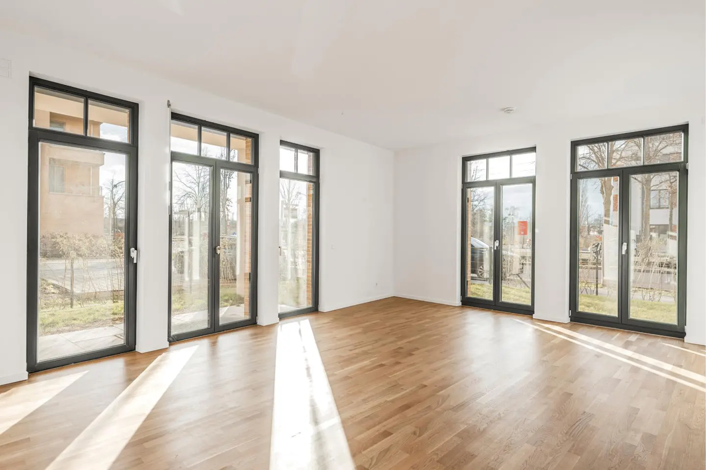 Bright, empty room with hardwood floors and white walls. Large black-framed windows offer outdoor views. Sunlight streams across the floor.