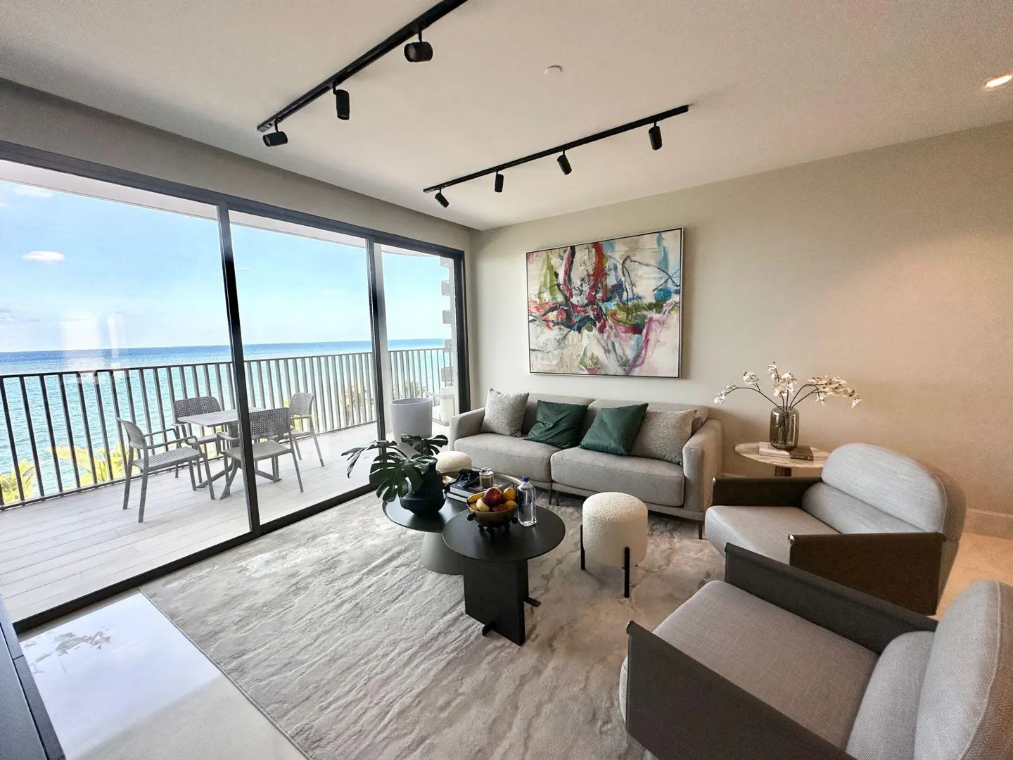 Living room with ocean view. Gray sofa, chairs, and rug. Black track lighting and coffee table. Abstract art on the wall. Balcony with table and chairs.