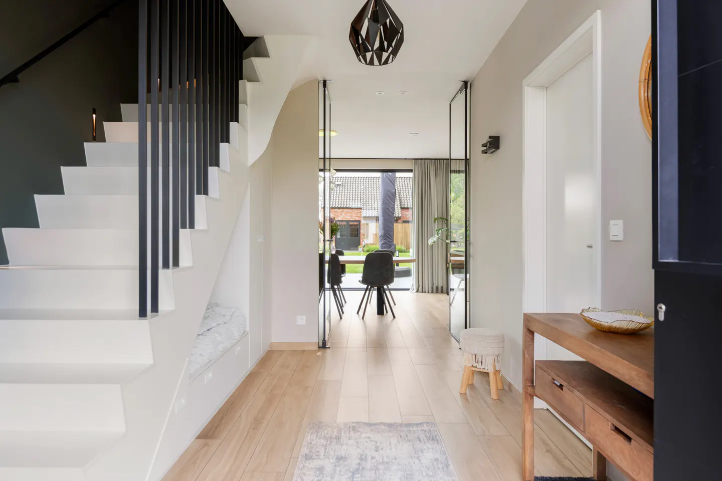 Bright foyer with white stairs and black railings. A wood floor leads to a glass door with a view of a dining table and backyard.