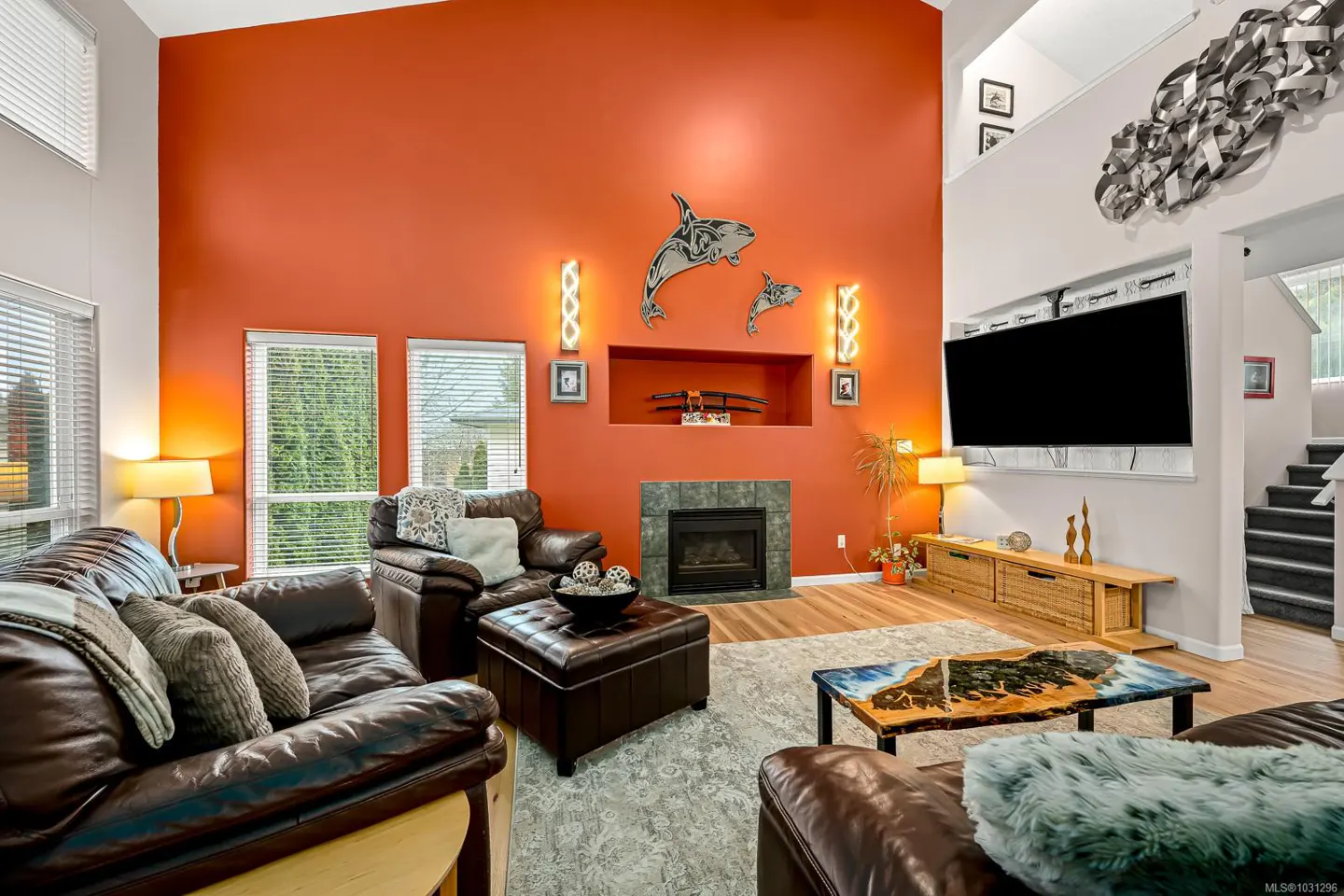 Living room with orange wall, brown leather sofas, fireplace, and large TV. Orca art and modern lighting add character. Windows provide natural light.