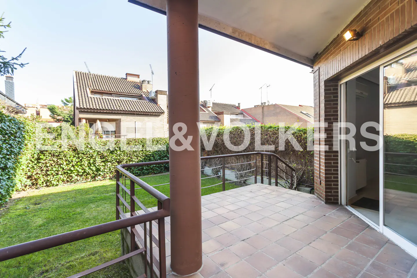 Covered patio with brick flooring, brown railings, and a view of a green lawn and houses in the background. Sliding glass doors to the right.