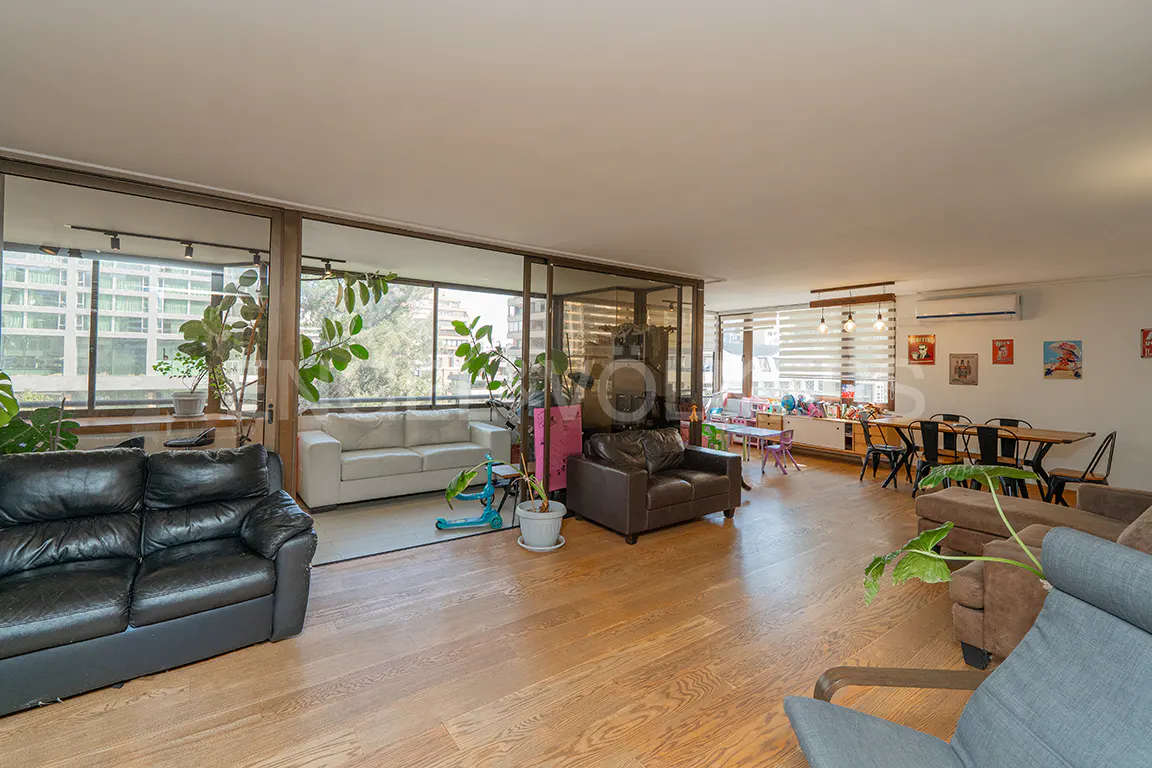 Bright, open living room with wood floors, sofas, dining table, and large windows with city views. Plants add a touch of nature.