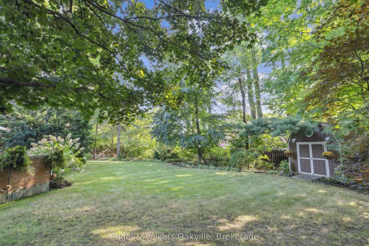 Lush green backyard with a shed. Trees surround the yard, providing shade. Flowers and plants add color.