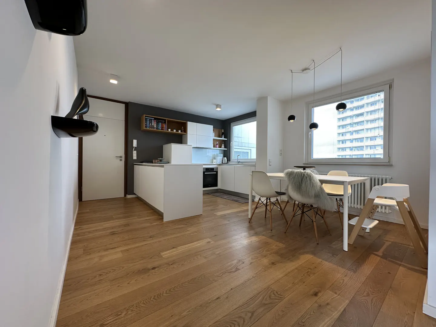 Bright, modern apartment interior with wood floors. White kitchen island and cabinets, dining table with chairs, and a large window.