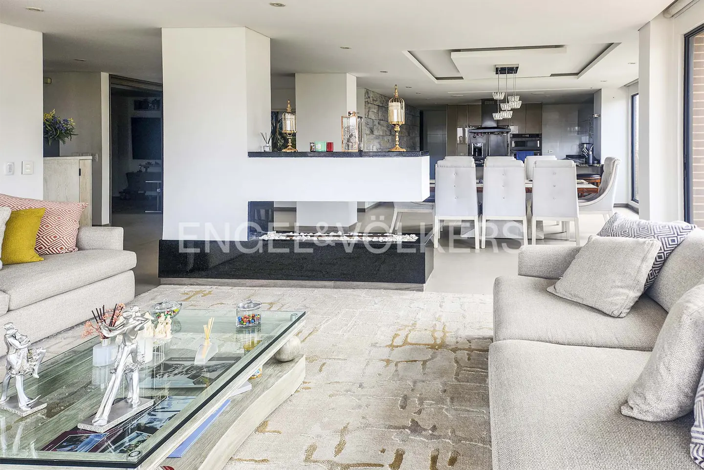 Modern open-plan living room with a central fireplace, glass coffee table, and dining area with white chairs.