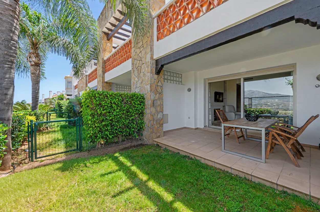 Exterior view of a condo with a patio, table, chairs, green lawn, and palm trees on a sunny day.