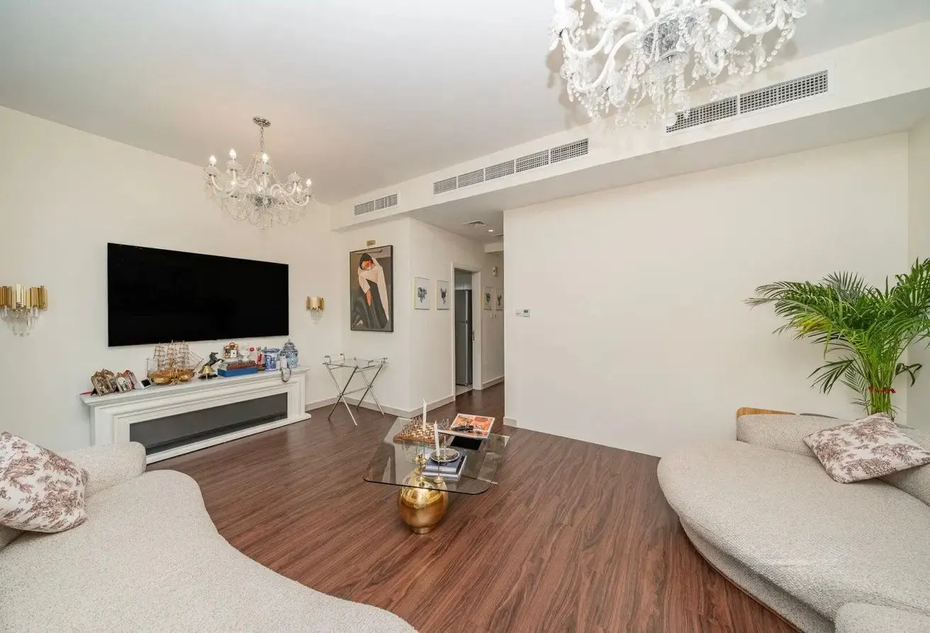 Bright living room with hardwood floors, white walls, and two chandeliers. A large TV sits above a white fireplace. Two curved sofas face each other.
