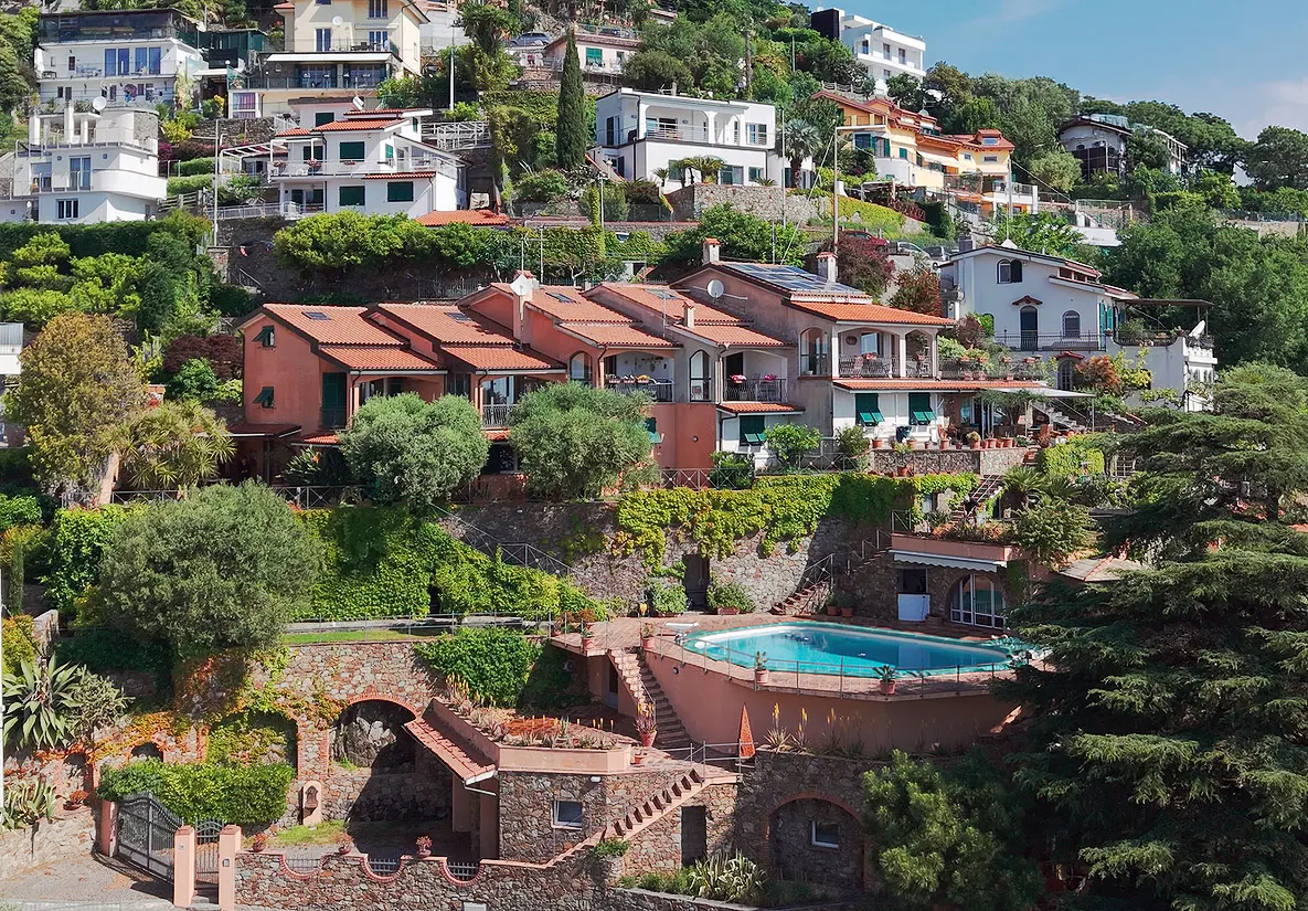 View of hillside homes with red tile roofs, lush greenery, and a blue swimming pool.