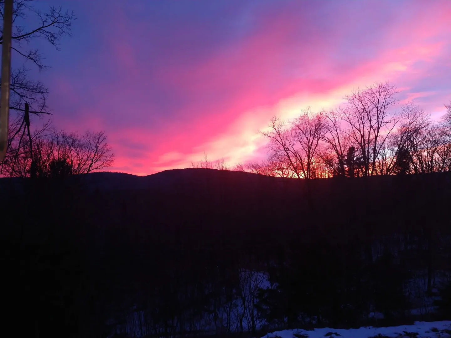 A vibrant pink and purple sunset over a dark, silhouetted treeline and distant hills.