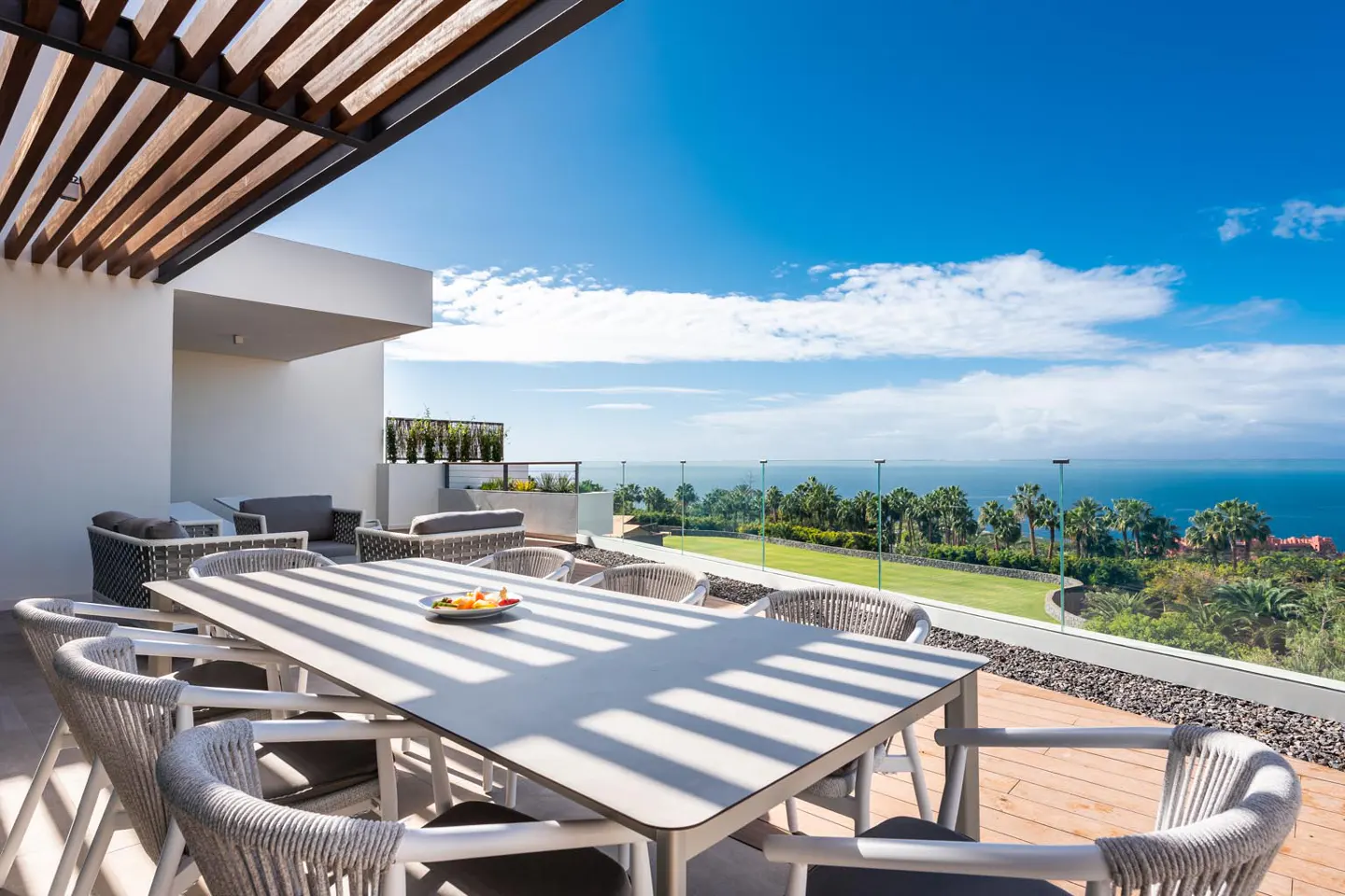 Outdoor patio with a white table and rope chairs under a pergola, overlooking the ocean and a blue sky.