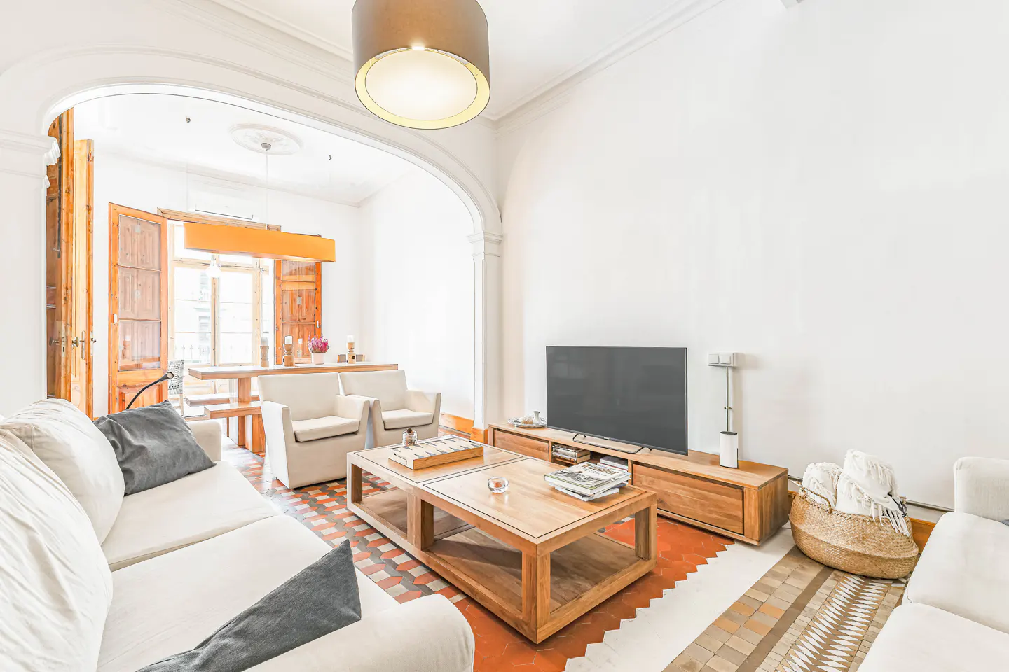 Bright living room with white sofas, wooden table, TV, and patterned tile floor. Arched doorway leads to dining area with open shutters.