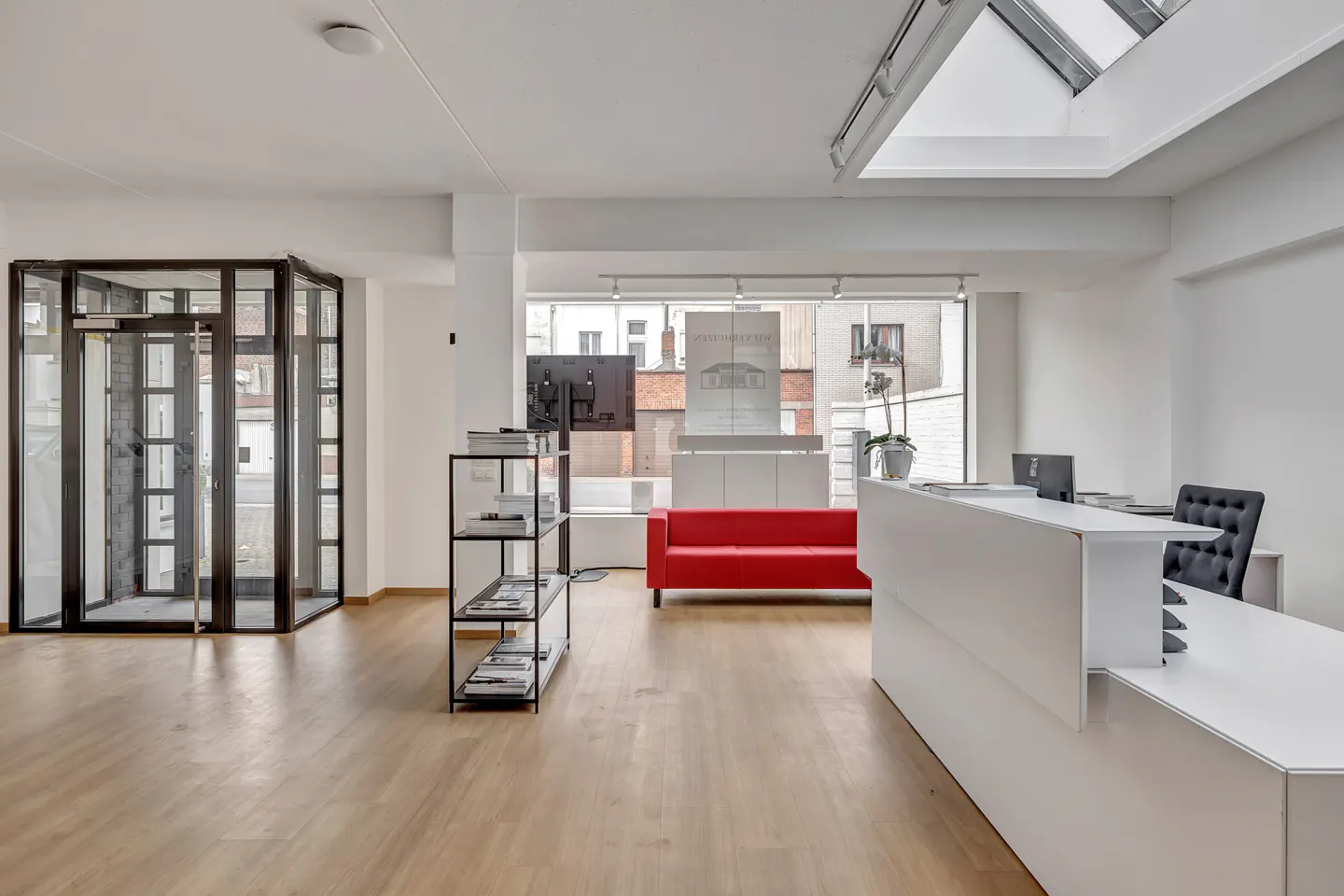 Bright real estate office with a white reception desk, red sofa, and black-framed glass doors. A bookshelf stands nearby.