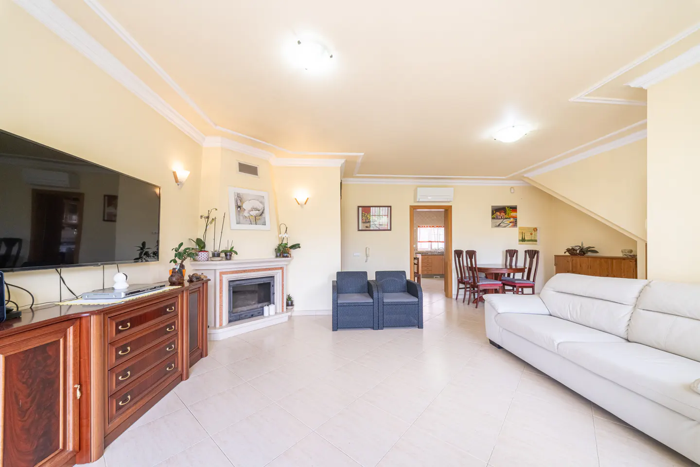 A bright living room with a white sofa, fireplace, TV, and dining table. The walls are yellow, and the floor is tiled.